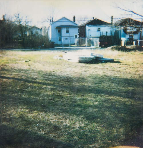 A grassy area with scattered debris, featuring two houses in the background and trees lining the edges.