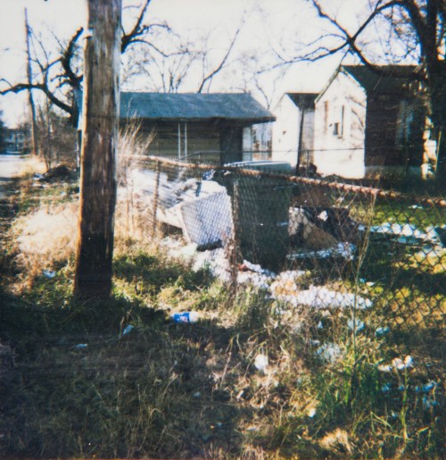 A rundown area with a fence, overgrown grass, and scattered debris near dilapidated buildings.