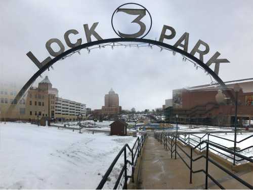 Archway sign for Lock 3 Park, with snow-covered ground and buildings in the background on a cloudy day.