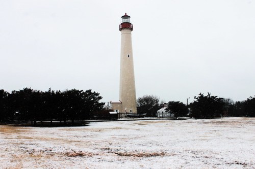 A tall lighthouse stands in a snowy landscape, surrounded by sparse trees and a field covered in light snow.