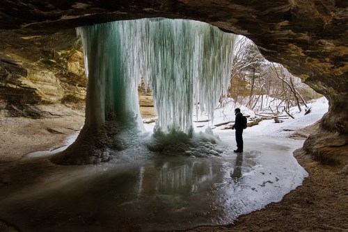 A person stands in a cave, gazing at a large frozen waterfall surrounded by icy terrain and rocky walls.