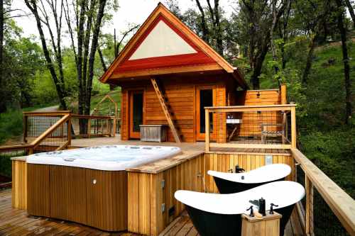 A wooden cabin with a hot tub and two bathtubs on a deck, surrounded by trees.