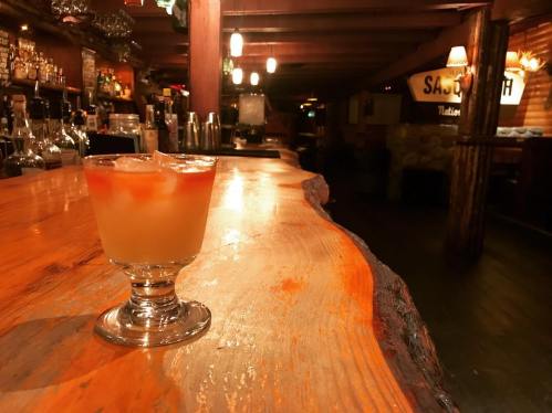 A cocktail sits on a wooden bar in a dimly lit tavern, with shelves of bottles in the background.