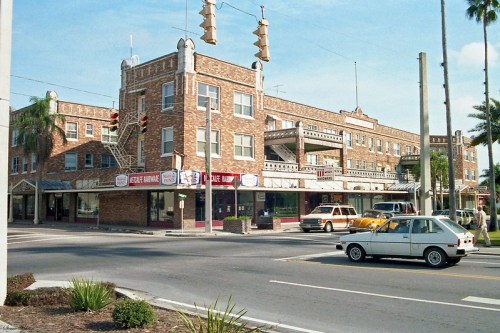 Historic brick building with balconies, shops on the ground floor, and palm trees lining the street. Classic cars parked nearby.
