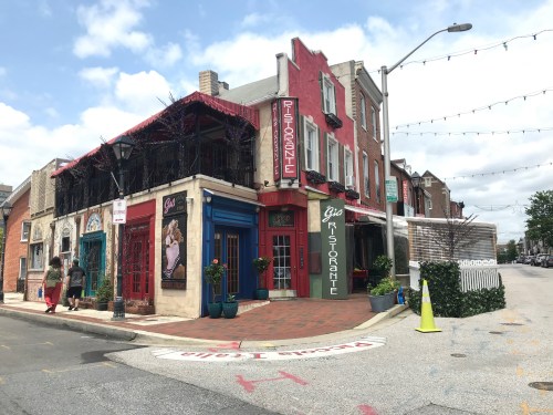 Colorful restaurant building at a street corner, featuring outdoor seating and decorative plants under a partly cloudy sky.