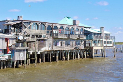 A waterfront building with wooden architecture and balconies, standing on stilts over calm water under a blue sky.