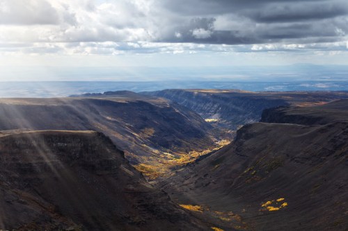 A dramatic landscape of deep canyons with autumn foliage, under a cloudy sky with rays of sunlight breaking through.