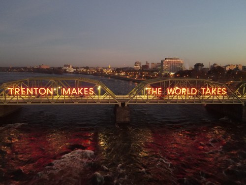 Neon sign on a bridge reads "Trenton Makes, The World Takes" with a city skyline in the background at dusk.