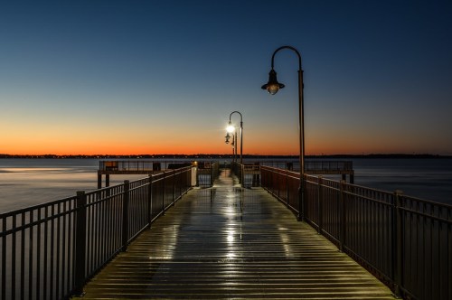 A serene pier at dusk, illuminated by street lamps, with calm water reflecting the twilight sky.
