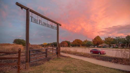 A sign for "The Flower Mound" at sunset, with colorful clouds and a red car parked nearby.