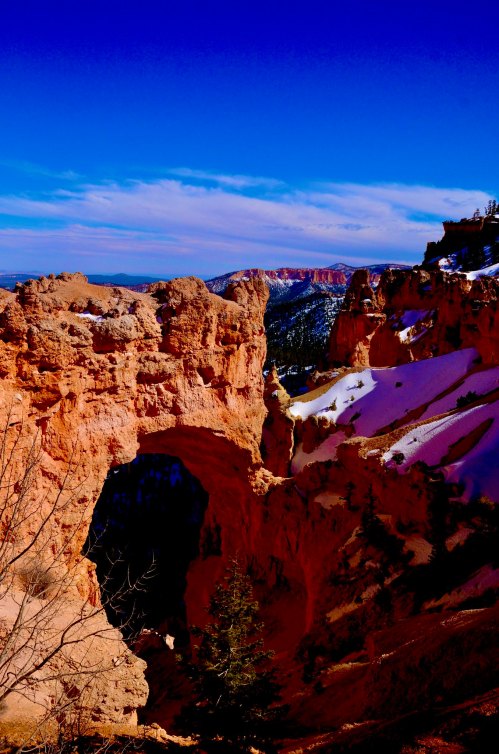 A stunning view of a natural rock arch surrounded by snow-covered cliffs and a clear blue sky.