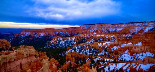 A panoramic view of snow-covered red rock formations under a dramatic blue sky at sunset.