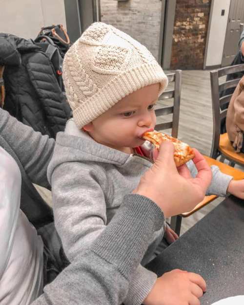 A toddler in a cozy hat enjoys a slice of pizza while being held by an adult.