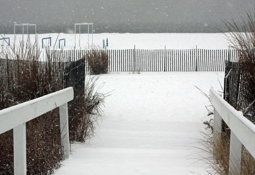 A snowy pathway leads to a beach, with a wooden fence and grass on either side, under a gray, overcast sky.