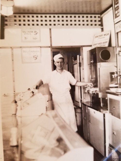 A vintage photo of a smiling man in a white uniform and cap, standing behind an ice cream counter.
