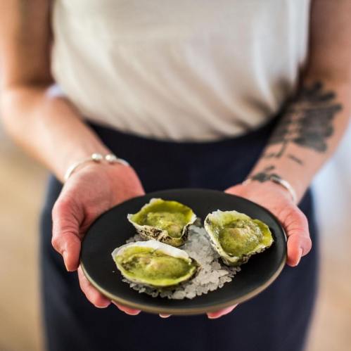 A person holds a black plate with three oysters topped with green sauce, resting on a bed of ice.