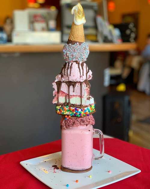 A tall, colorful dessert stack featuring ice cream, a cone, and toppings, served in a glass with a pink milkshake.