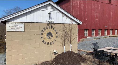A rustic market building with a sign reading "Homestead Market," surrounded by gravel and a red barn in the background.
