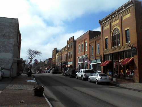 A quaint street lined with historic brick buildings, shops, and parked cars under a partly cloudy sky.