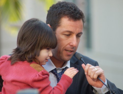 A man in a suit holds a young child while speaking into a microphone at an outdoor event.