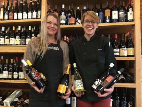 Two women smile while holding bottles of wine in a store, surrounded by shelves filled with various wine bottles.