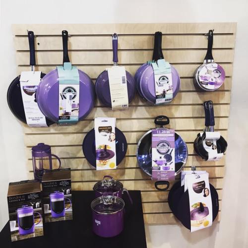 Display of purple cookware, including frying pans, pots, and a French press, arranged on a wooden wall.