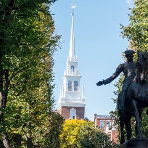 A tall church steeple rises above trees, with a statue of a horseman in the foreground against a clear blue sky.