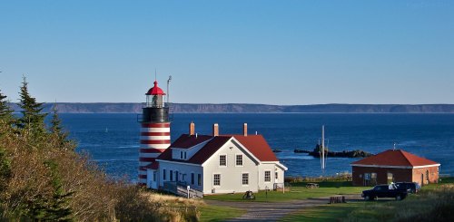A red and white striped lighthouse beside a white building, overlooking the ocean and distant hills under a clear blue sky.