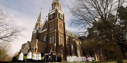 A large church with a tall clock tower, surrounded by people in white attire, set against a clear sky.