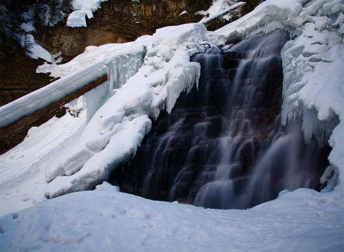 A frozen waterfall surrounded by snow and ice, cascading down rocky terrain in a winter landscape.