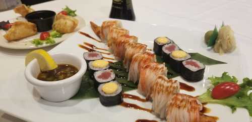 A plate of sushi featuring rolls with shrimp, avocado, and dipping sauce, alongside fried appetizers and garnishes.