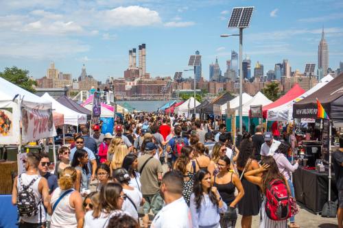A bustling outdoor market with crowds, food stalls, and a skyline view of New York City in the background.