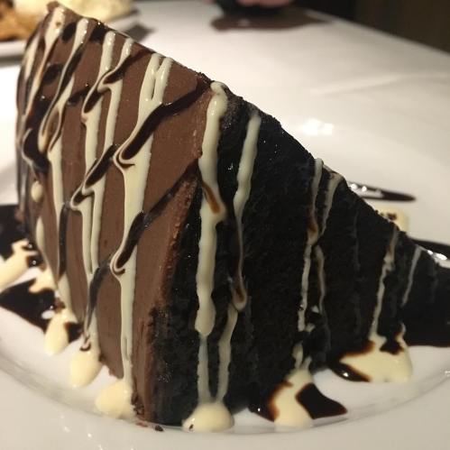 A slice of rich chocolate cake drizzled with chocolate and cream sauces on a white plate.