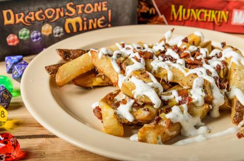 A plate of loaded fries topped with bacon and drizzled with sauce, with board games in the background.
