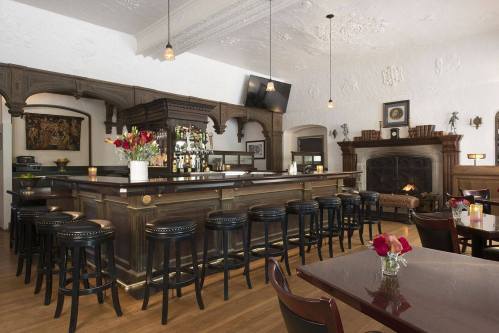 A cozy bar with a wooden counter, high stools, and a fireplace, adorned with flowers and elegant decor.