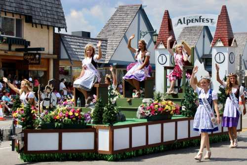 A festive parade float featuring women in traditional dresses waving, surrounded by flowers and decorative scenery.