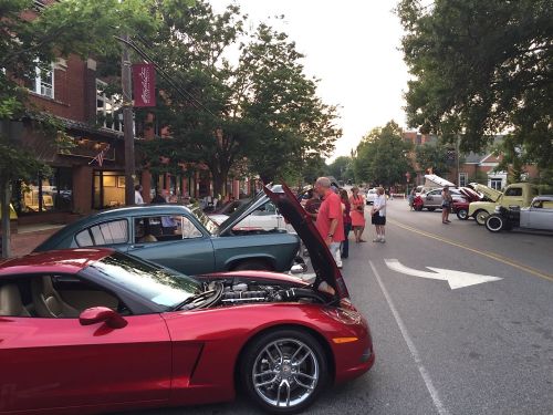 A street lined with classic cars and spectators, with a red sports car in the foreground and trees in the background.