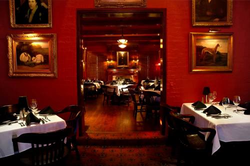 Elegant restaurant interior with red walls, framed paintings, and tables set with white tablecloths and black napkins.