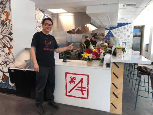 A person stands next to a counter with flowers, wearing a black shirt with a logo, in a modern kitchen setting.