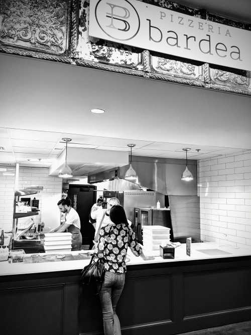 A woman stands at the counter of Pizzeria Bardea, watching staff prepare food in a modern, stylish setting.