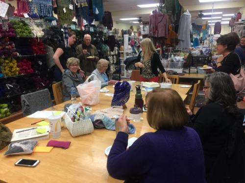 A group of people gather around a table in a yarn shop, enjoying snacks and engaging in conversation while crafting.