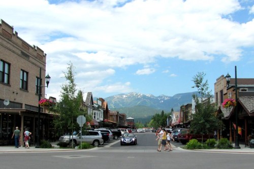 A charming street lined with shops and trees, with mountains in the background under a partly cloudy sky.