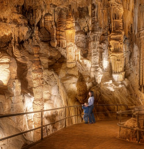 A child and an adult explore a cave with illuminated stalactites and stalagmites, surrounded by rocky formations.