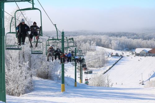 Skiers and snowboarders on a chairlift, surrounded by snowy hills and frosted trees on a clear winter day.