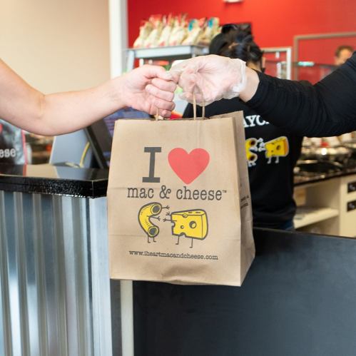 A person hands over a takeout bag with "I ❤️ mac & cheese" printed on it at a restaurant counter.