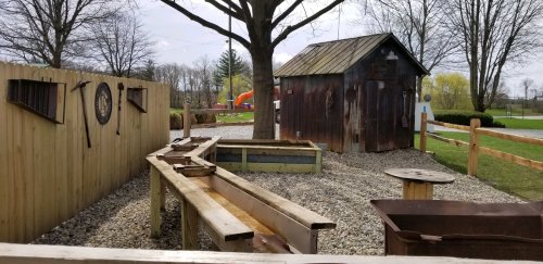 A rustic outdoor area with a wooden structure, gravel, and a small shed, surrounded by trees and playground equipment.