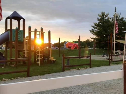 Sunset behind a playground with slides and climbing structures, surrounded by trees and festive lights.