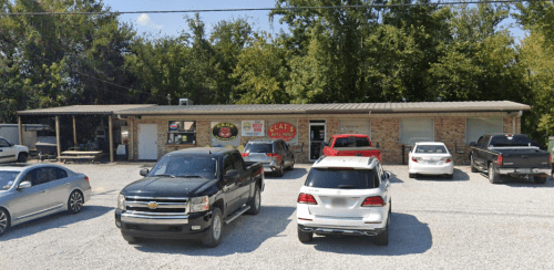 A small brick building with a sign reading "Clay's" and several parked cars in a gravel lot surrounded by trees.
