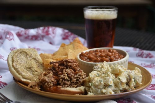 A plate with a pulled pork sandwich, potato salad, baked beans, tortilla chips, and a glass of beer.