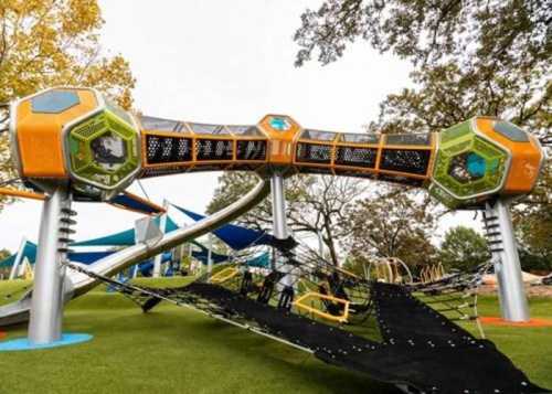 A modern playground structure with colorful hexagonal features and a netted climbing area on green grass.